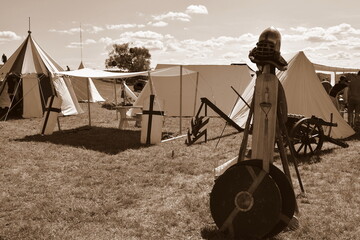 A sepia photo of a medieval camp full of cloth tents, stands, targes, shields, and knight equipment...