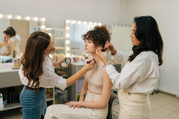 Side view of professional makeup artist applying foundation brush on neck body of young woman model. Hairdresser doing stylish hairdo to curly haired female. Concept of four-handed makeup and styling.
