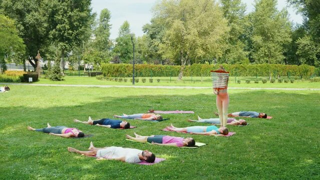 Yogi woman playing swinging chimes during meditation while practitioners lie on grass in corpse pose in city park. Yoga class deep restoration, body and mind relaxation concept