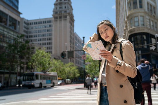 confused asian woman traveler pushing hair back and studying route map with concentration on the street while visiting downtown san Francisco in California usa on breezy day