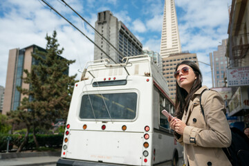 asian chinese female commuter wearing sunglasses looking into distance while expecting her bus to come in san Francisco's Chinatown. a white electric city bus is stopping at background