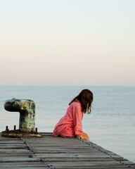 A girl, in a long pink shirt, greets the morning on a pier in the ocean, thinking about life.