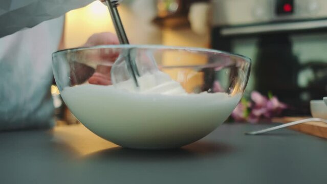 Women's hands beat egg whites in a glass bowl into a fluffy foam. Close-up of the table. Cooking in the kitchen