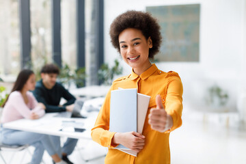 Portrait of happy black lady student showing thumb up, standing in classroom with classmates...