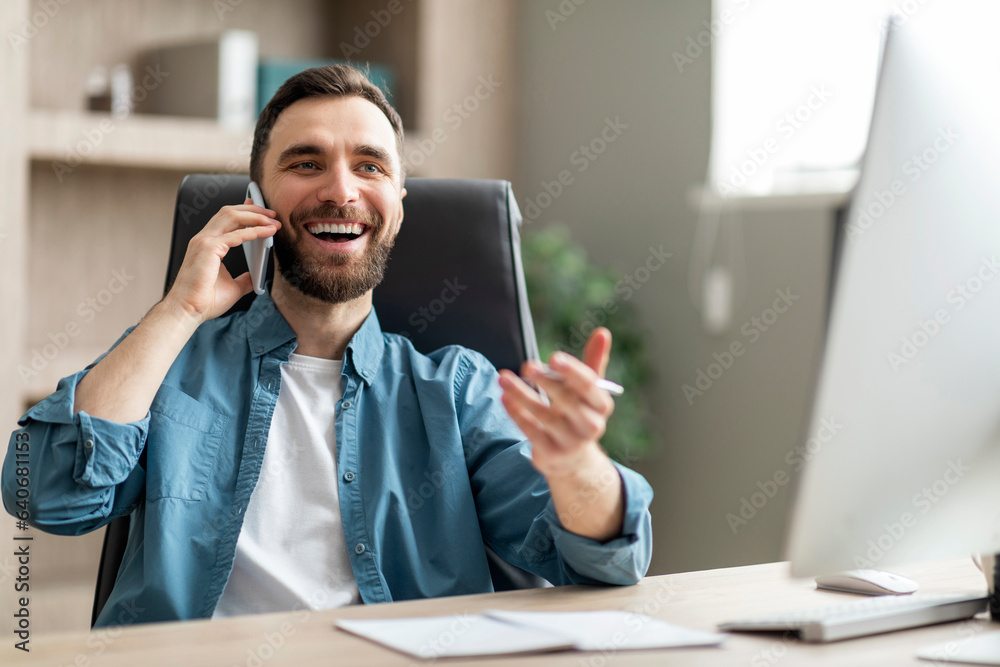 Poster smiling businessman talking on cellphone while sitting at desk in modern office