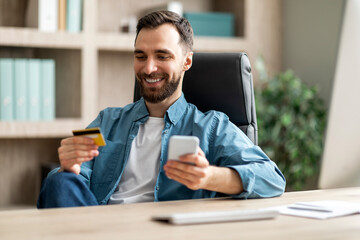 Online Payment. Smiling Young Businessman Using Smartphone And Credit Card In Office