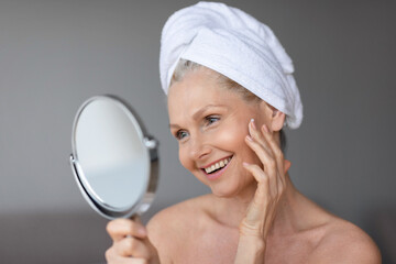 Facial treatment concept. Beautiful middle aged woman in towel on head looking in mirror and touching her perfect skin