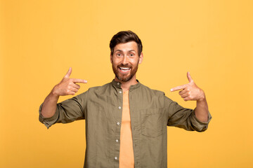 Happy middle aged man pointing fingers at himself and smiling to camera standing over yellow studio background