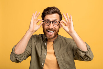 Excited middle aged man touching his glasses and smiling at camera, standing on yellow studio...