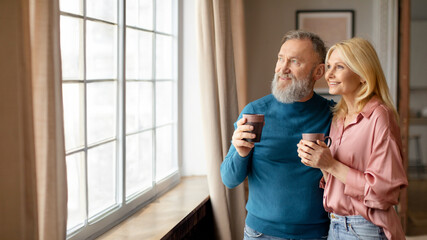 Happy Senior Couple Drinking Coffee Hugging Near Window At Home