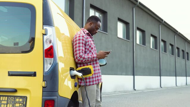 African american man chatting on mobile phone while electric car is charging on outdoor station