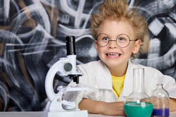 Joyful child with hair on end is engaged in science. An enthusiastic cheerful boy in lab coat poses...