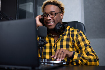 Young African man in glasses and a plaid shirt laughs while making a podcast in his home studio. A black man wearing headphones records the broadcast using a professional microphone and a DJ console