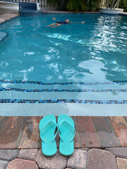 Woman floating in an outdoor luxurious swimming pool leaving her thong sandals on the edge.