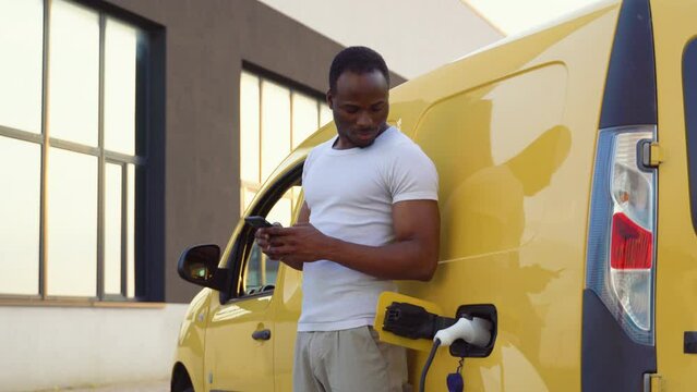 Black man chatting on mobile phone while electric car is charging on outdoor station
