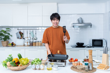 Happy portrait of asian young man of sit a cheerful preparing food and enjoy cook cooking with vegetables, while standing on a kitchen Condo life or home