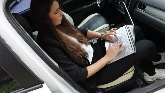 Close-up of a business lady working on her laptop sitting in her car. Beautiful girl model in black jacket. High quality video