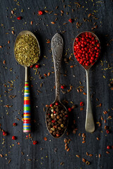 Spoons with oregano, red and mixed peppercorn on black background