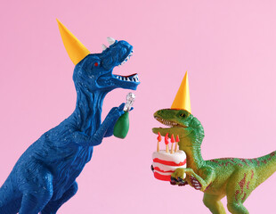 Cute plastic toy blue and green dinosaurs in party hats with cake and champagne bottle on pink...