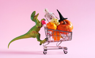 Happy green dinosaur with shopping trolley full of Halloween decorations on pink background.