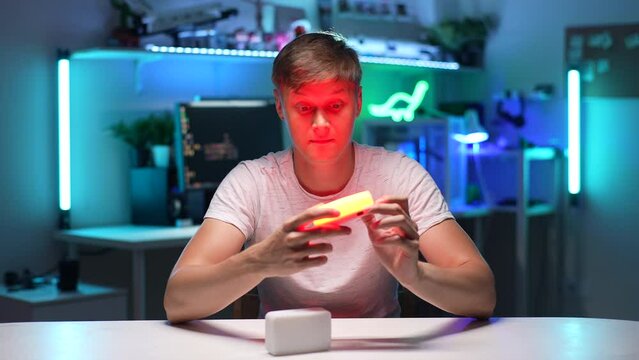 Portrait of videographer male looking, examining, testing out professional new mini LED lamps sitting at desk on background of desktop computer with neon light background. Shooting in slow motion.