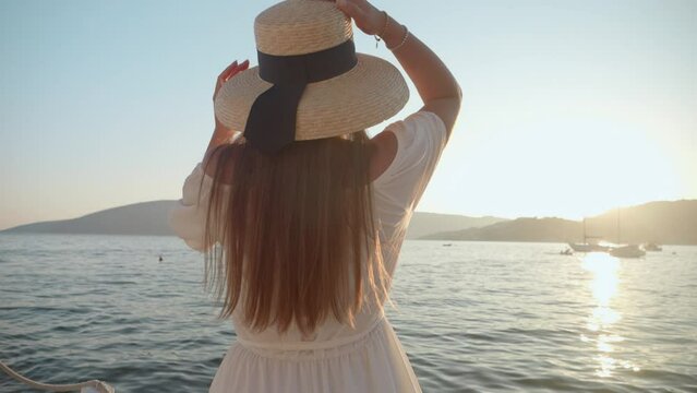 A young brunette with long hair, donned in a white dress and hat, runs in slow motion on a wooden pier by the sea. Tourism, summertime, holiday, and beachside vacation.