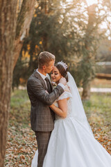 A fabulous romantic newlywed couple is hugging at sunset in an autumn park with leaves from trees on the grass
