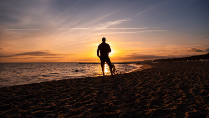 A man standing in front of the sunrise at the Baltic Sea. Next to the man is a camera on a tripod. Sandy beach in Kolobřeh. Kołobrzeg is city in Poland. - obrazy, fototapety, plakaty