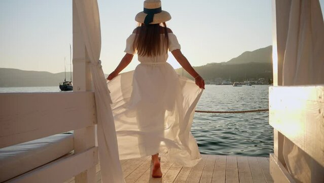 Slow motion of young brunette girl in a white dress, hat, and flowing hair running and dancing on a wooden pier by the sea . Tourism, summertime, holiday, and beach vacation.