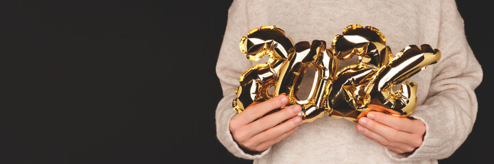 2024 - New Year concept. Banner with woman in sweater hold in hands golden inflatable foil balloons in front of black background. - Powered by Adobe