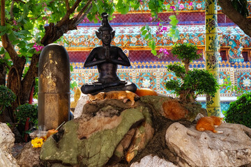 A statue of a meditating figure in a garden setting. The statue is black and is sitting on a rock formation with orange cats sleeping on it. The background also has trees and plants.