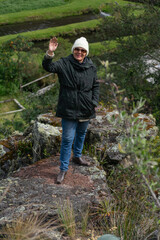 Senior Latin hiker waving her hand and smiling at camera while standing on a rock. Active and healthy lifestyle.