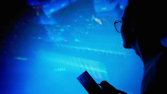 A woman wearing glasses in complete darkness looks at charts and graphs of the Forex market. Screensaver. Free space for insertion.