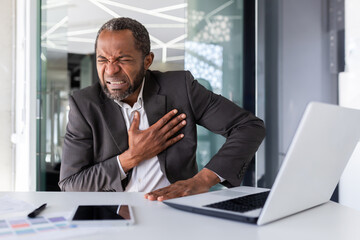 Naklejka premium Heart attack in mature experienced man inside office at workplace, senior african american holding hands on chest, pain is severe in boss in business suit.
