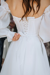 The bride in a white dress ties a corset ribbon with a bow on her back by hand. Close-up wedding photography, portrait.