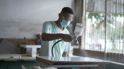 One young black worker applying paint spray on wooden surface at carpentry workshop - obrazy, fototapety, plakaty