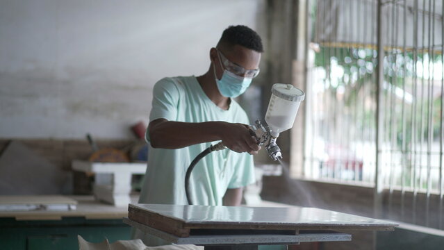 One young black worker applying paint spray on wooden surface at carpentry workshop