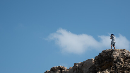 Unrecognized photographer takes photos with camera  on rocky peak. Blue cloudy sky above the cliff