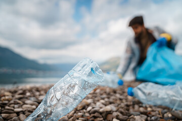 Closeup selective focus photo of plastic bottle stuck on the beach and unrecognizable obscured mindful man cleaning garbage and recyclables in the background.