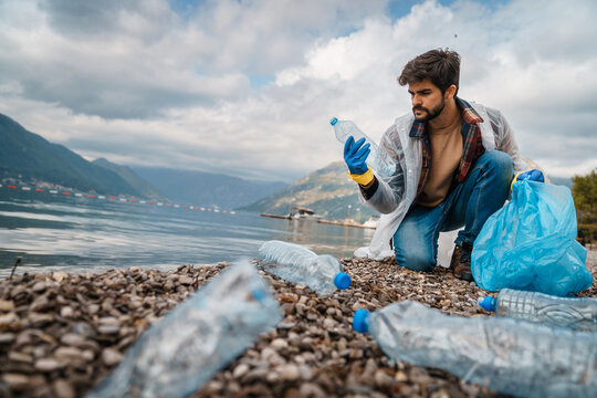Concentrated serious unselfish man wearing protective gloves, protecting water life from pollution, picking up the garbage and recyclables from the beach.