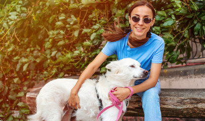 Happy young pretty brunette girl hugging her cute white fluffy dog on the outdoor in the park on lawn. Adoption, rescued, shelter, companion, pet.