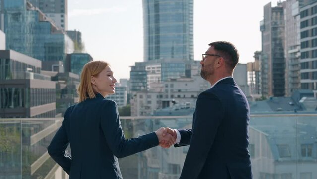 Back view handheld shot two successful business partners male female handshaking deal gesture celebrate success walk along office building roof terrace overlooking high-rise skyscrapers on sunny day