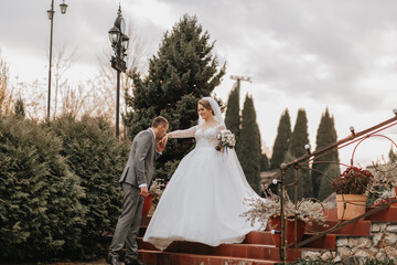 Fototapeta premium Stylish bride and groom tenderly embrace and kiss. Beautiful bride with modern bouquet embracing stylish groom in autumn park. Sensual romantic moment. Wedding. Stylish and beautiful.