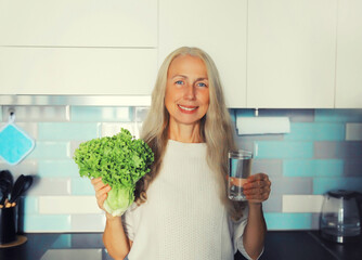 Portrait of happy smiling middle-aged gray-haired woman holds green lettuce and glass with fresh...