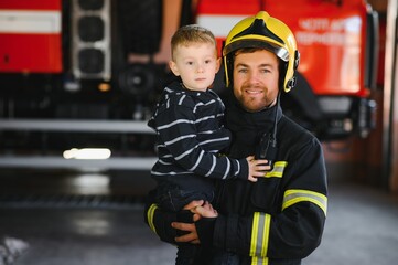 A firefighter take a little child boy to save him. Fire engine car on background. Fireman with kid in his arms. Protection concept.