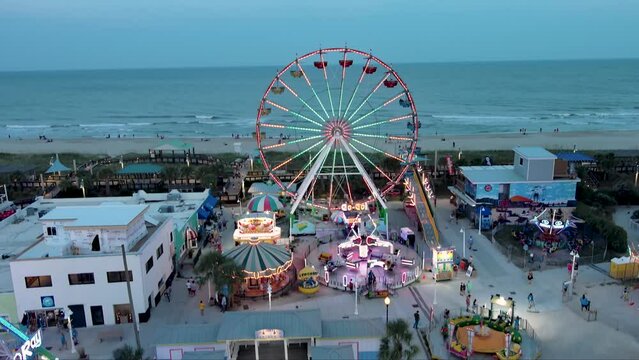 ascending aerial footage of the Carolina Beach Boardwalk with a Ferris wheel, colorful amusement rides, people walking, a sandy beach, blue ocean, hotels and condos at sunset in Carolina Beach
