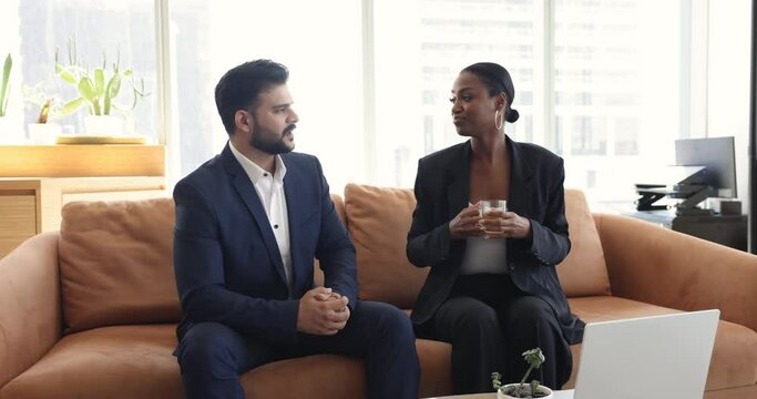 Two young multiracial colleagues, African and Indian mates in formal suits spend break seated on sofa in modern office talking drinking tea, share ideas discuss joint project. Teamwork, companionship