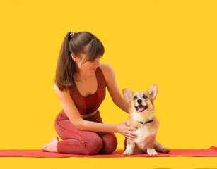 Sporty young woman sitting on yoga mat with cute Corgi dog against yellow background