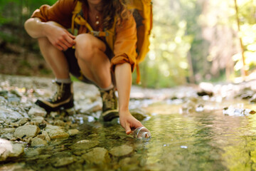 Woman traveler with a hiking yellow backpack explores new places, collects water from a stream, climbs up the mountains along a forest hiking trail.