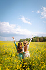 Girlfriends dressed in stylish dresses are walking together in a rapeseed field. Two women enjoy...